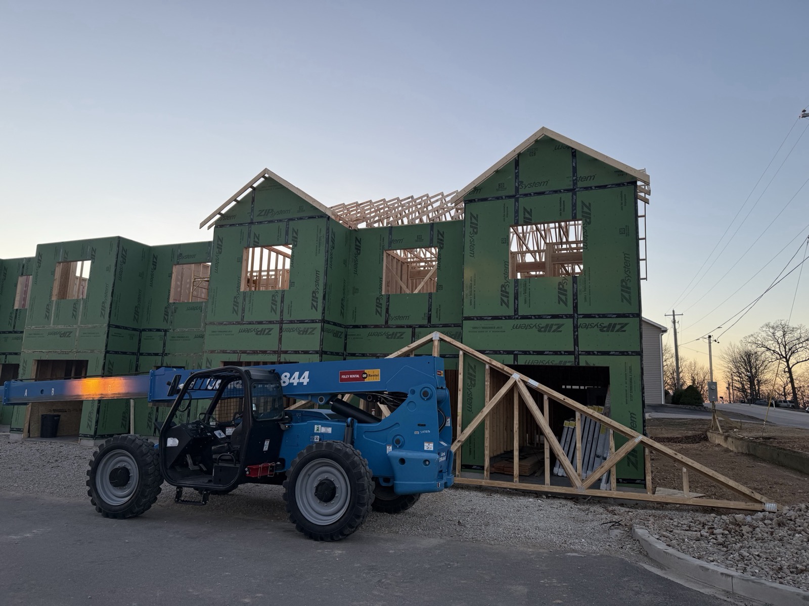 Two-story building framed in weather sheathing at dusk with roof trusses staged and a telehandler on site, an Ireland Construction project